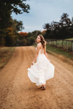 A figure in a flowing white dress twirls and dances along a rustic dirt road surrounded by autumn trees.