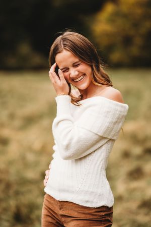 A person in a cozy white sweater and brown pants smiles joyfully while posing in an autumn field at golden hour.