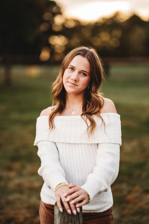 A woman in a cozy white sweater poses outdoors during golden hour with soft autumn bokeh in the background.