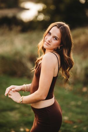 A woman in a brown dress poses outdoors during golden hour with soft natural lighting in the background.