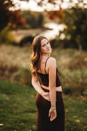 A person in a black dress poses outdoors during golden hour with autumn foliage in the background.