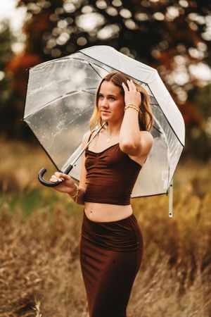 A figure poses with a clear umbrella in a brown outfit against a golden autumn field during sunset.