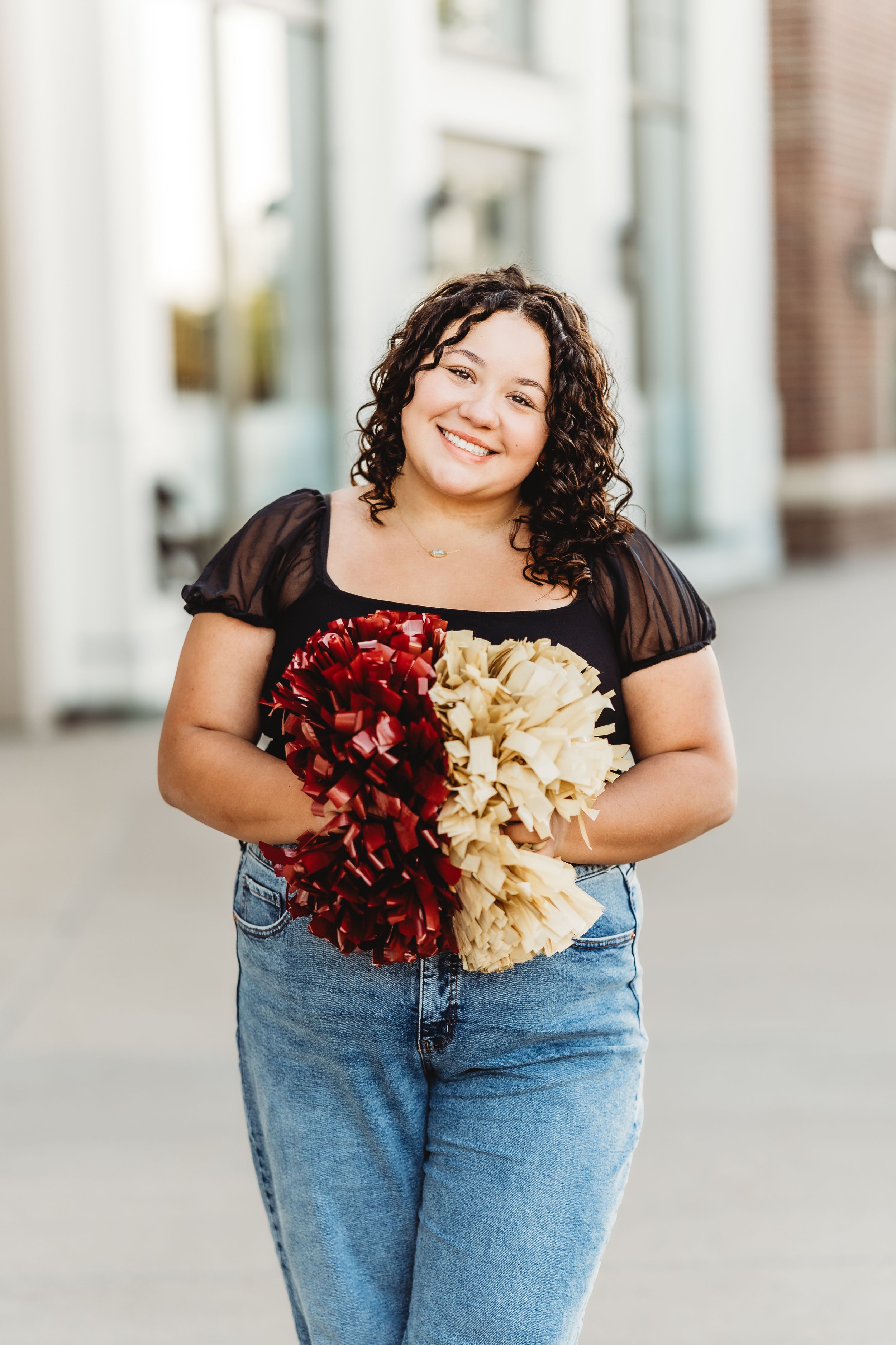 Cheerleader in denim jeans and black top poses with red and gold pom poms against a light urban backdrop.