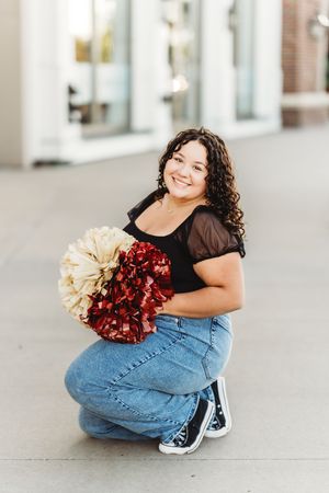 A cheerleader in jeans and black top poses with red and gold pom poms on a sidewalk.