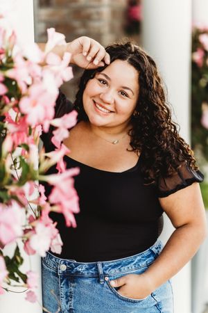 Cheerleader in casual attire poses near pink flowers and white columns in an outdoor setting.