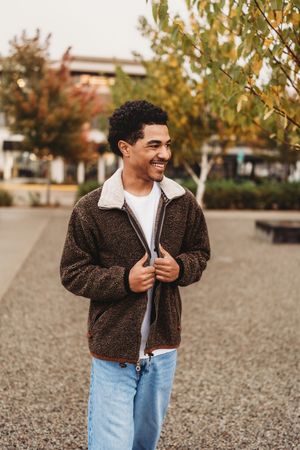 A person in a brown fleece jacket and light jeans stands outdoors on gravel during autumn.