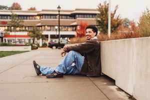 A person sits casually on a concrete ledge wearing jeans and a brown jacket on an autumn college campus.