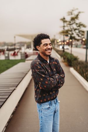 A person in a brown patterned sweater and jeans poses joyfully on a walkway by the ocean on an overcast day.