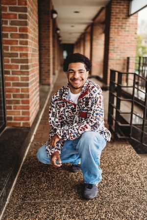 A person in a patterned shirt and jeans poses casually in a brick-lined walkway with gravel flooring.