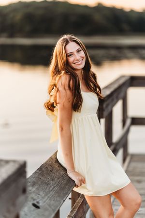A person in a white top poses playfully on a wooden dock at sunset near a lake or river.