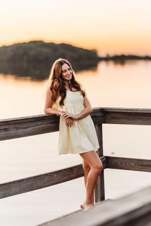 A person in a white summer dress poses on a wooden dock at sunset against a serene waterfront backdrop.