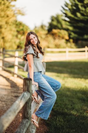 A person in blue jeans and a gray top sits casually on a wooden fence post in a sunny outdoor setting.