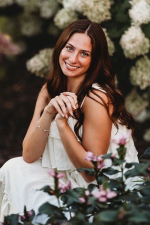 A woman in a white dress poses gracefully among blooming white flowers and green foliage in an outdoor portrait series.