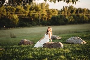 A person in a flowing white dress reclines on grassy hills at golden hour as sunlight filters through trees.