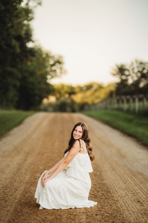 A person in a flowing white dress poses gracefully on a sunlit dirt road surrounded by green trees and grass.
