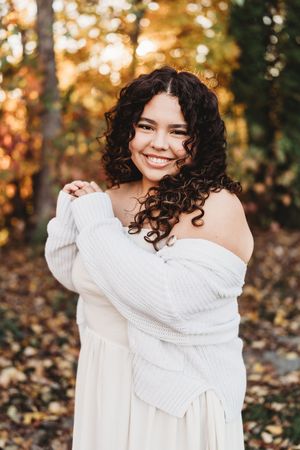 Woman in white sweater poses outdoors in autumn setting with golden bokeh and fallen leaves in background.