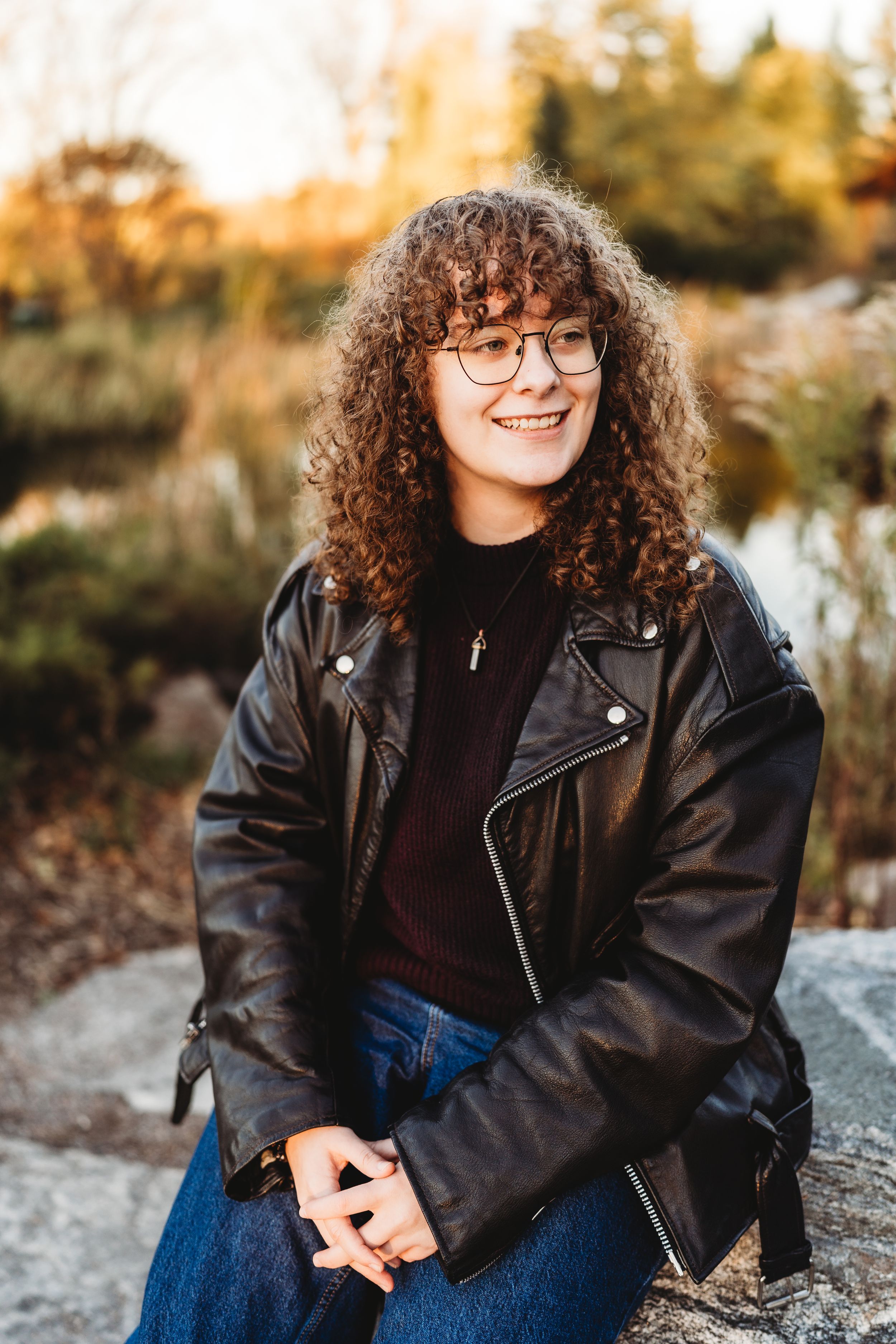 Person wearing a black leather jacket and glasses sits casually outdoors during golden hour sunset.