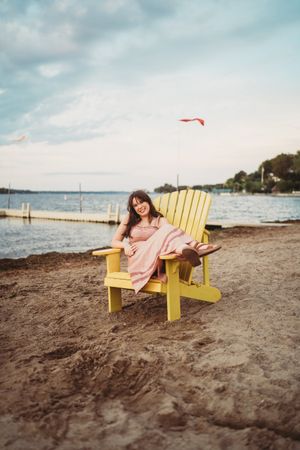 A person relaxes in a yellow Adirondack chair on a sandy beach with cloudy skies and water in the background.