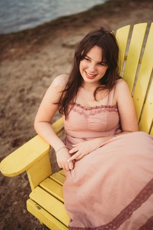 A person in a peach-colored dress relaxes in a bright yellow Adirondack chair on a beach.