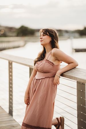 Someone wearing a dusty rose sundress poses along a waterfront railing at sunset.