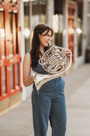 A person in striped overalls holds a shiny French horn instrument against a blurred urban background.