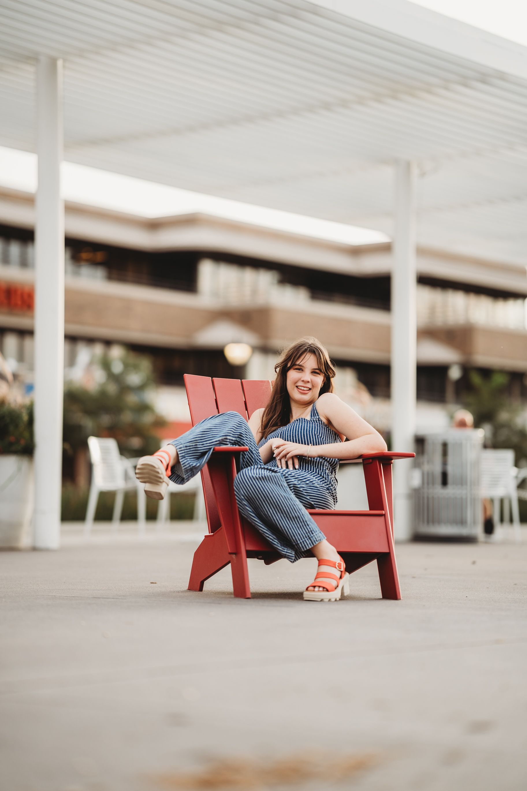 A person relaxes in a red Adirondack chair wearing jeans and sandals on a covered outdoor patio.