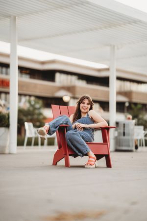 A person relaxes in a red Adirondack chair wearing jeans and sandals on a covered outdoor patio.