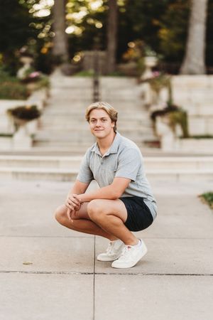 A person in a grey polo shirt and shorts crouches on concrete steps outdoors on a sunny day.