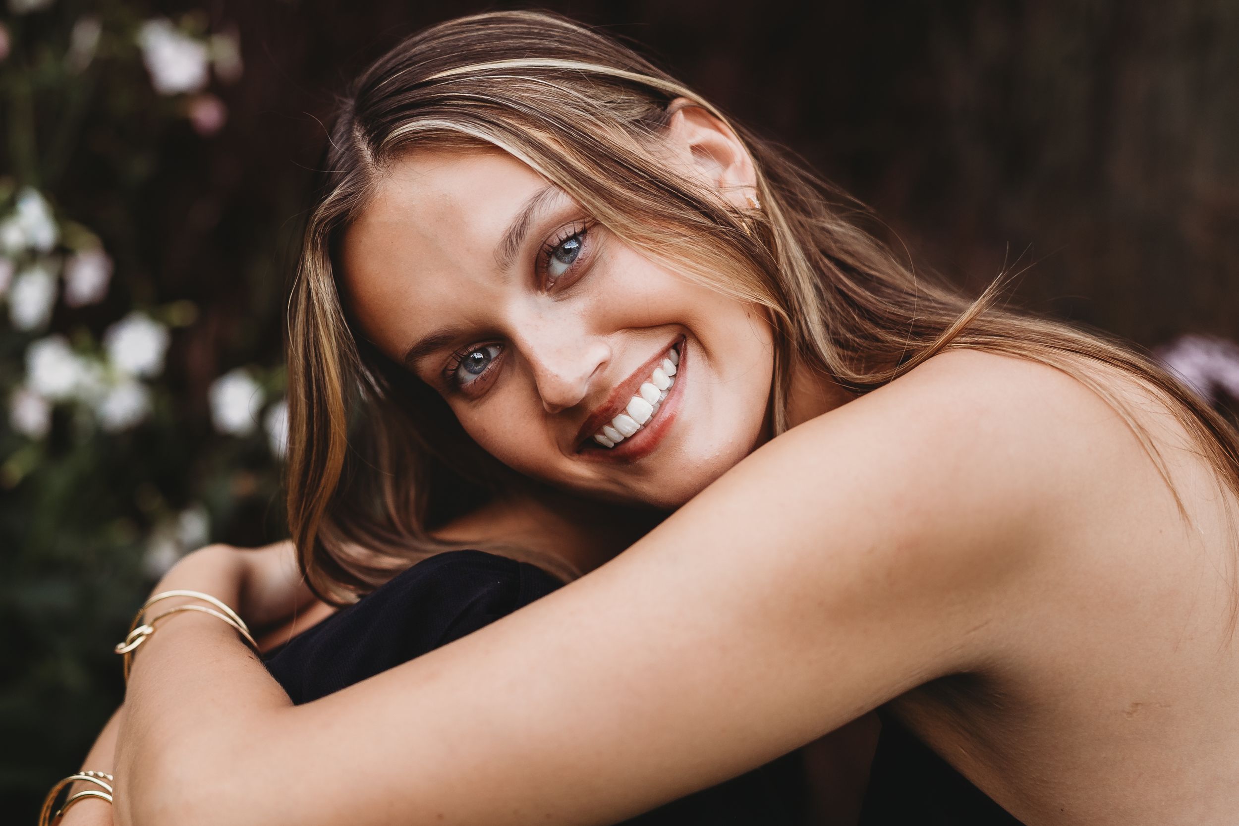 A smiling person with long blonde hair poses outdoors in natural lighting against a dark background.
