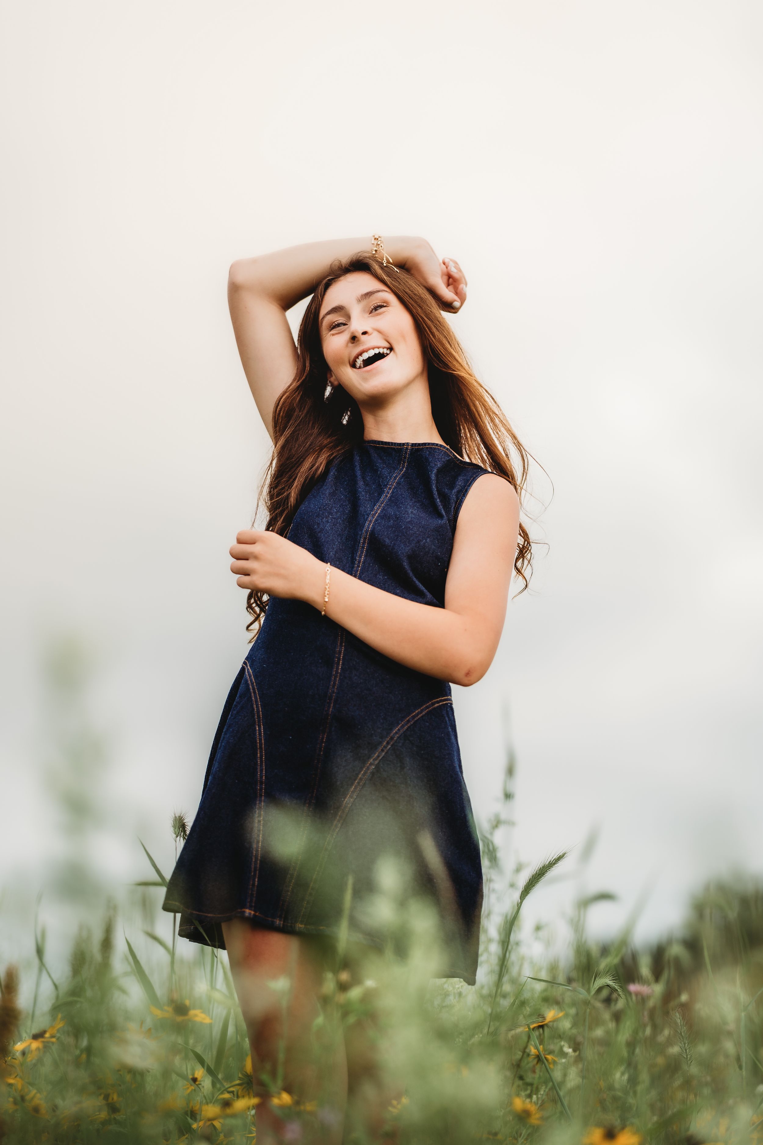 Happy young woman in navy denim dress laughing in wildflower field with arm raised overhead.