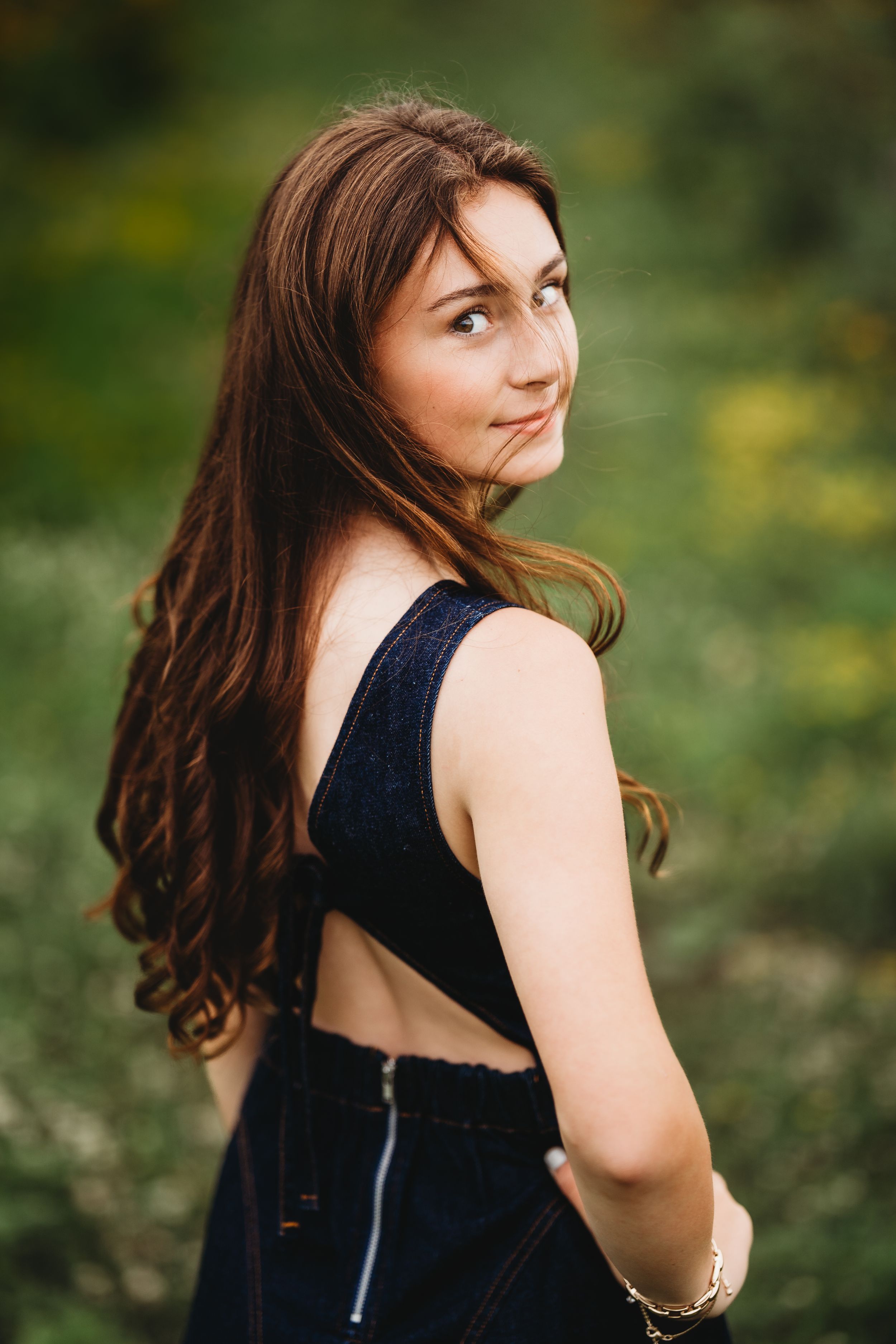 Young woman with long brown hair in a black backless outfit poses outdoors in a green field, glancing over shoulder.