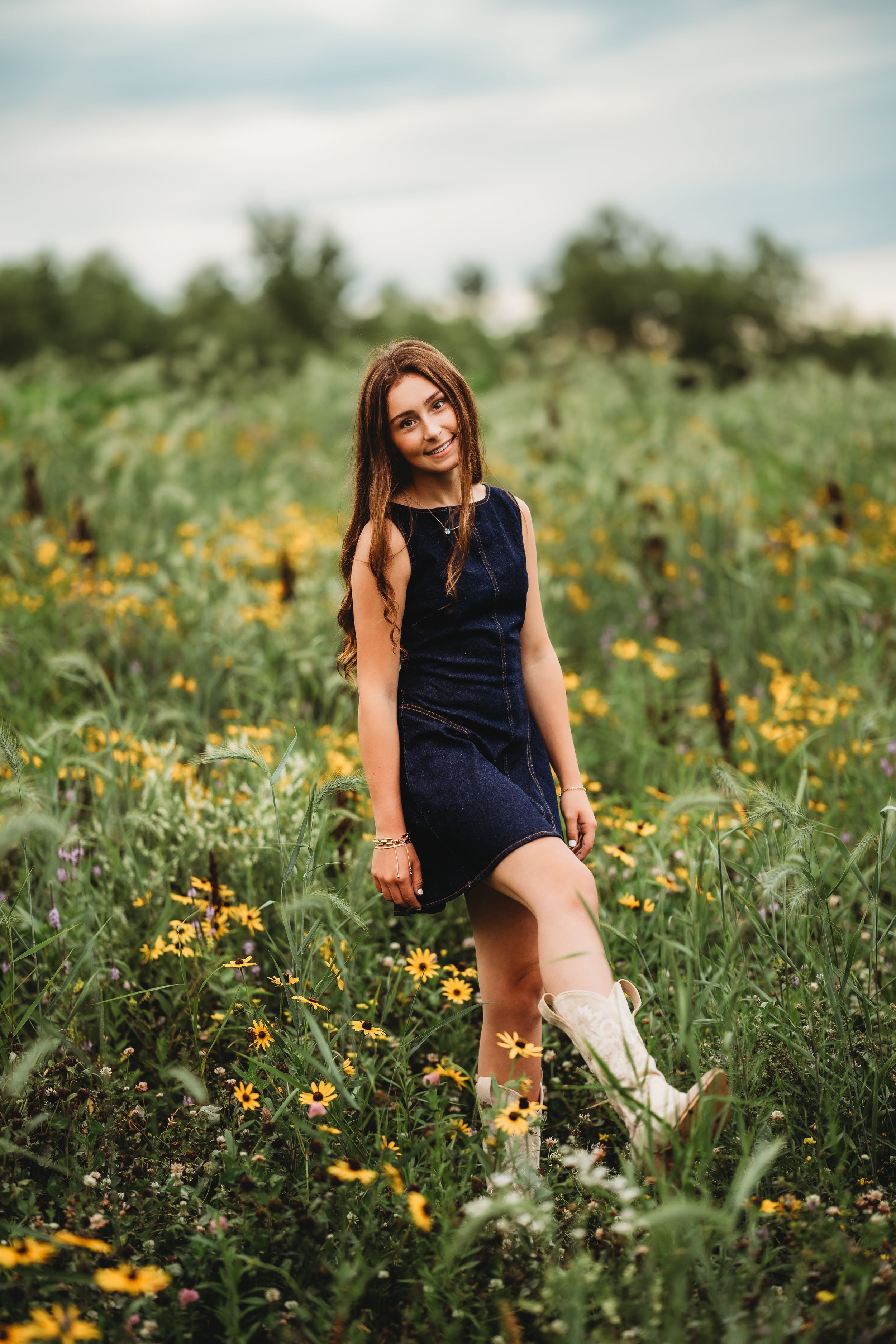 Young woman in navy dress and white boots posing in a wildflower field with yellow blooms.