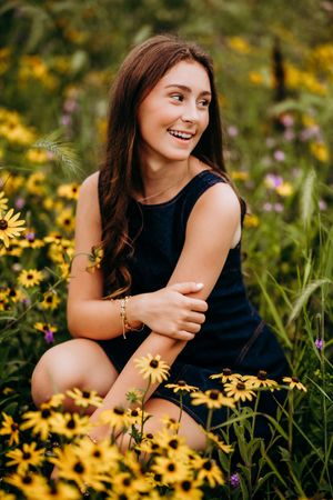 Smiling young woman in navy dress sitting among yellow black-eyed Susan wildflowers in a lush green field.