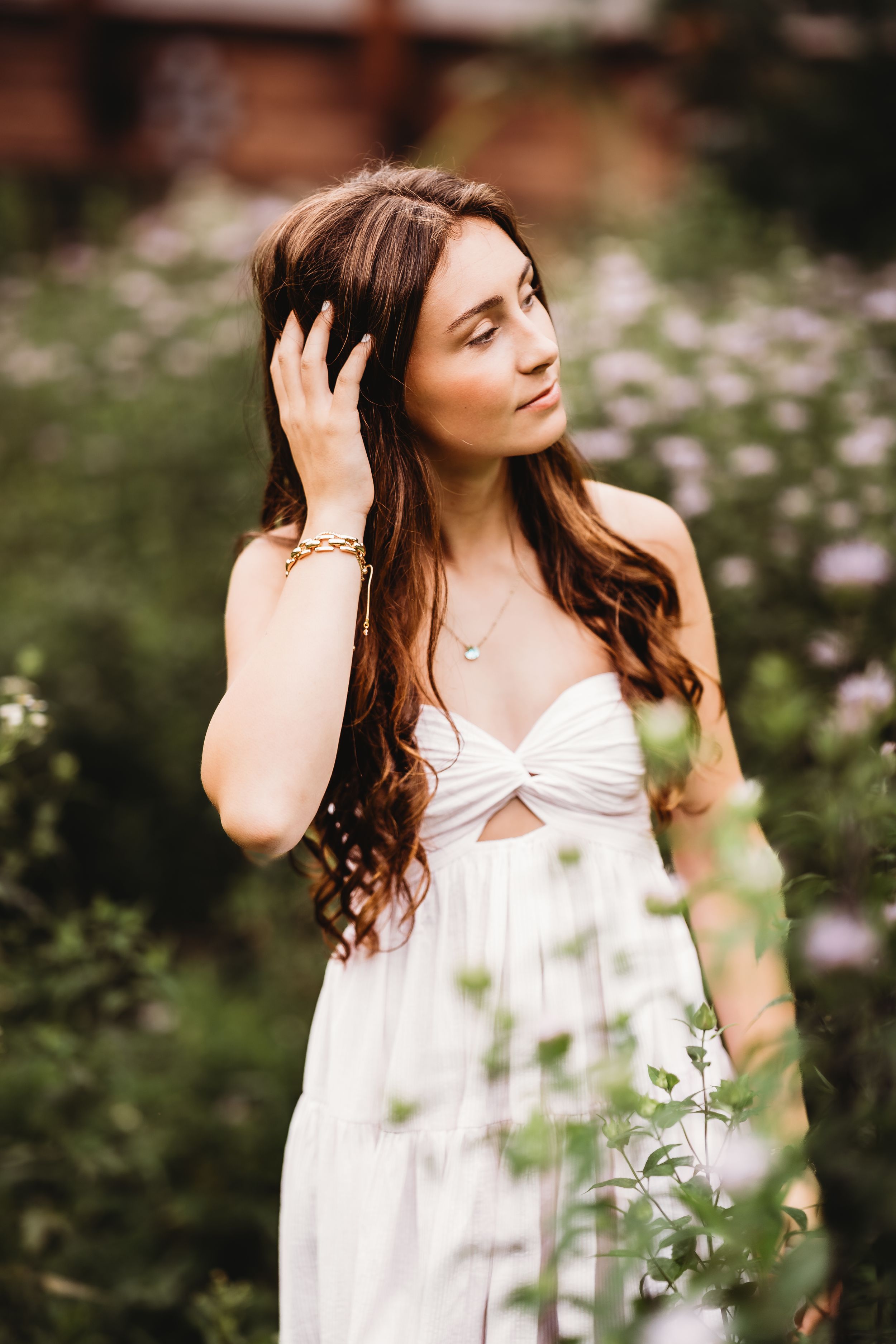 Young woman in white strapless dress poses among wildflowers, touching her hair, wearing gold bracelet and necklace.