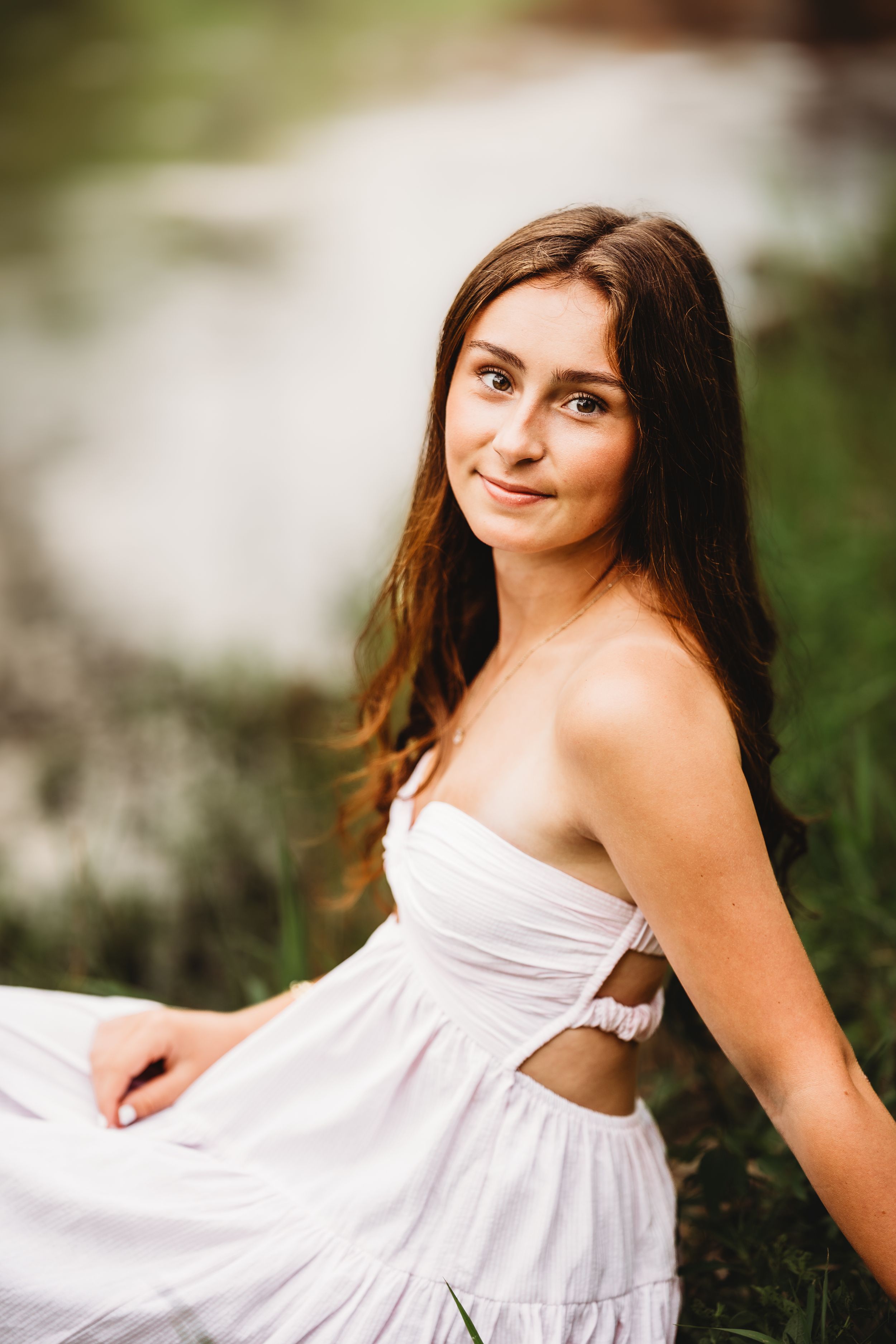 Young woman in white sundress sitting in green grass near water, smiling softly at camera.