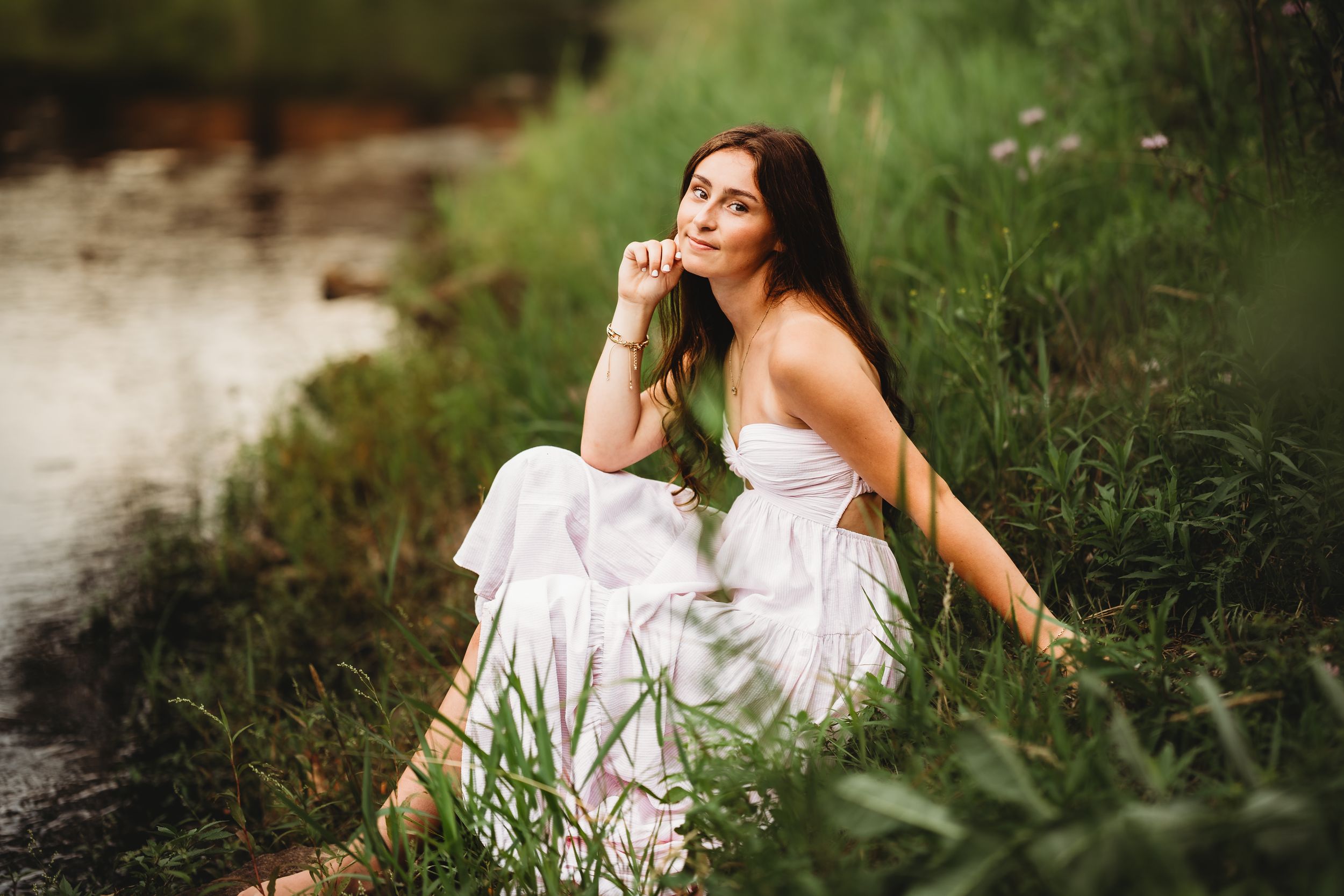 Young woman in white dress sitting by a peaceful stream surrounded by lush green grass and wildflowers.