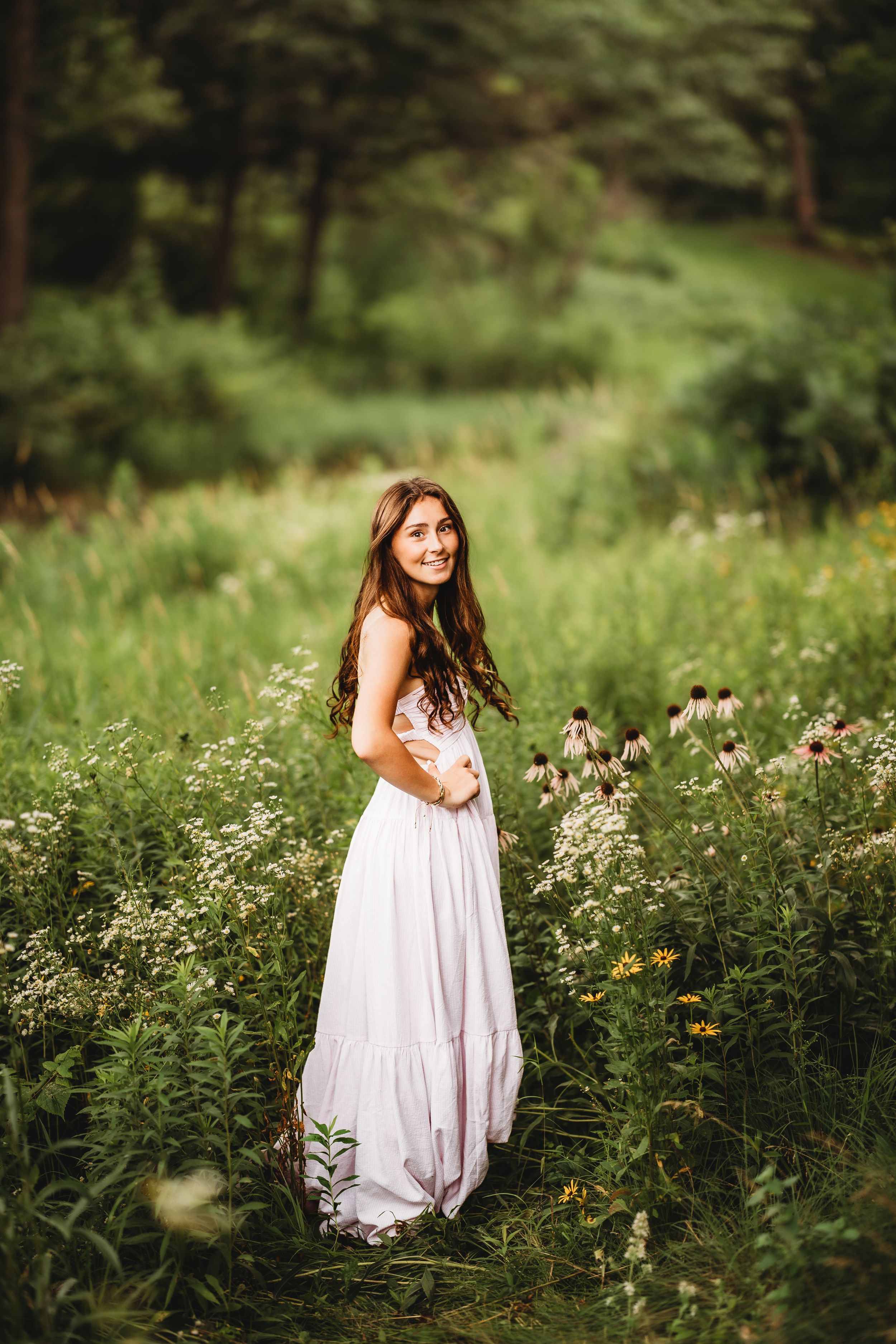Young woman in white maxi dress smiling in a wildflower meadow surrounded by lush green trees.