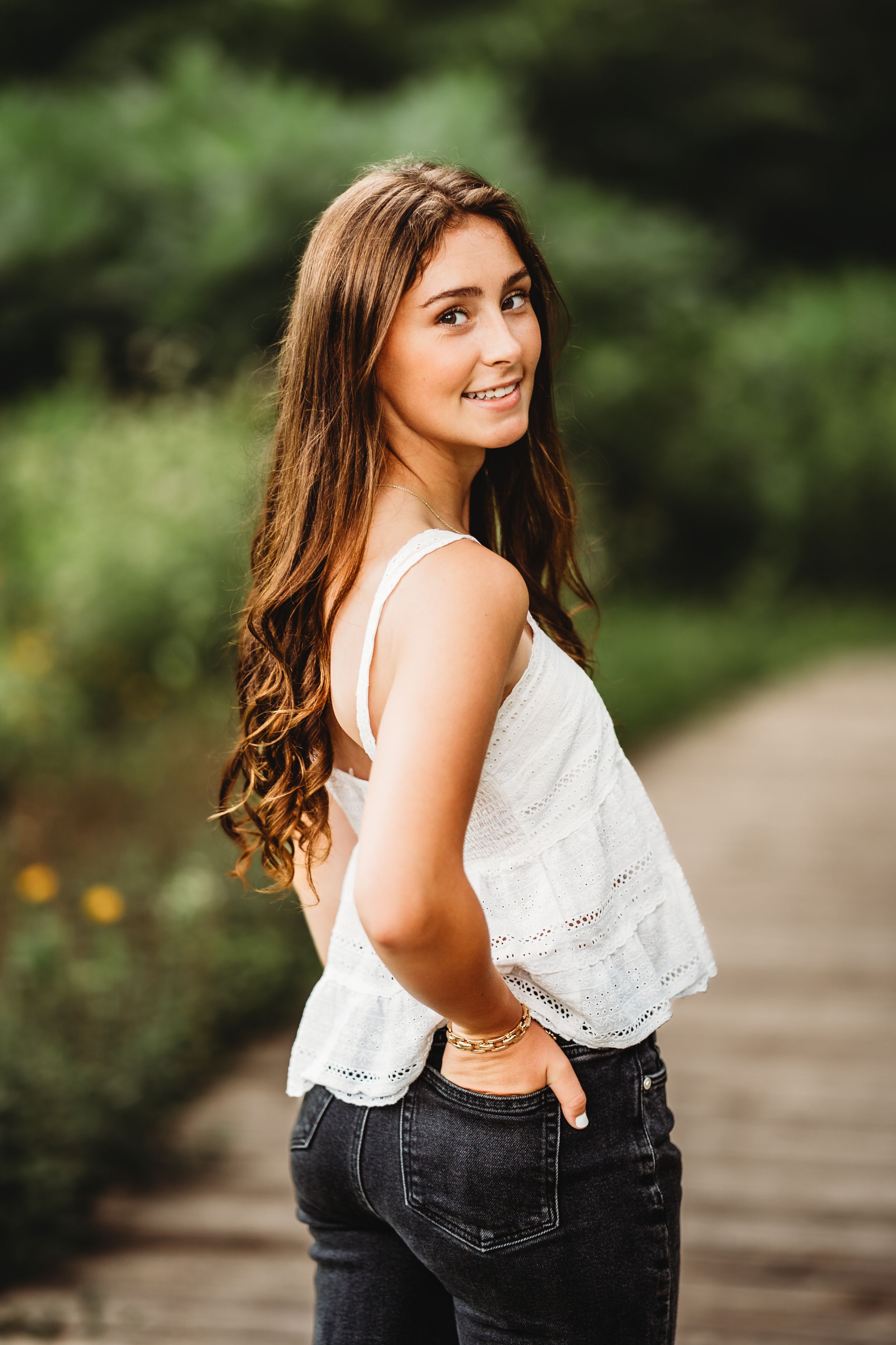 Young woman with long brown hair smiles over shoulder, wearing white top and dark jeans on a wooden path outdoors.