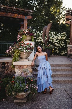 A woman in a light blue tiered ruffle dress poses on stone steps near decorative garden planters at sunset.