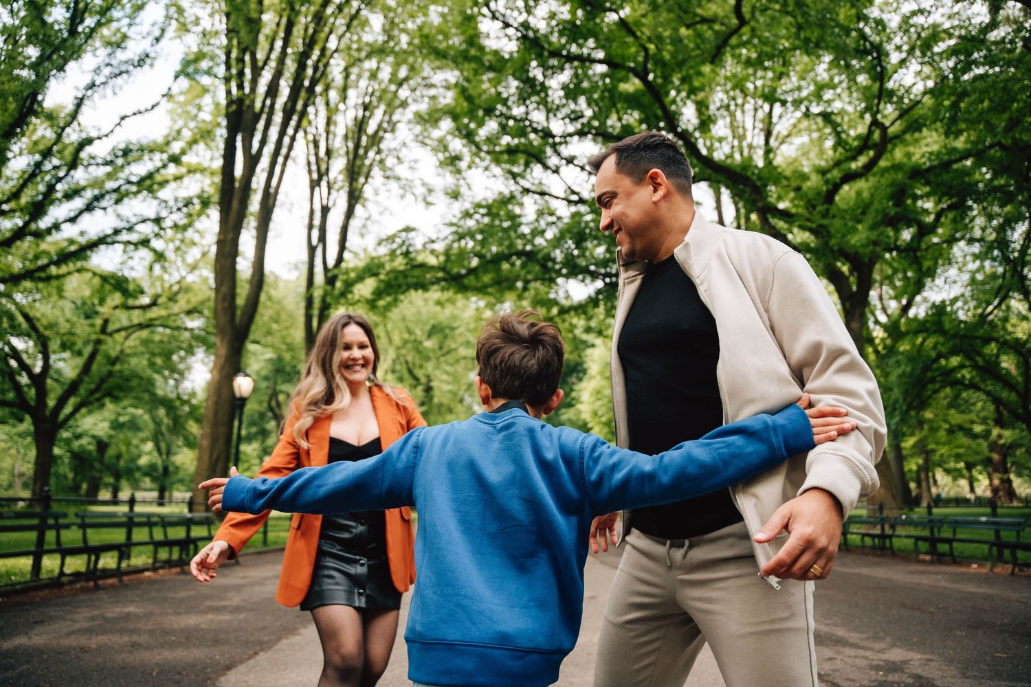 A family walks together on a tree-lined park path with outstretched arms on an autumn afternoon.