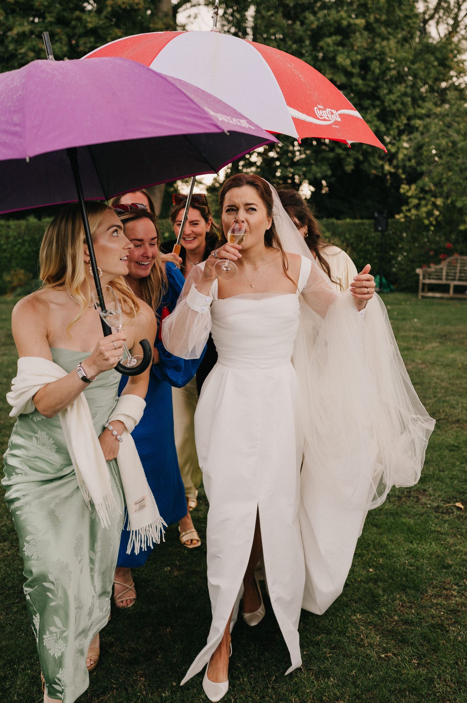 Wedding party laughing together under umbrellas on a rainy day in a green outdoor setting.
