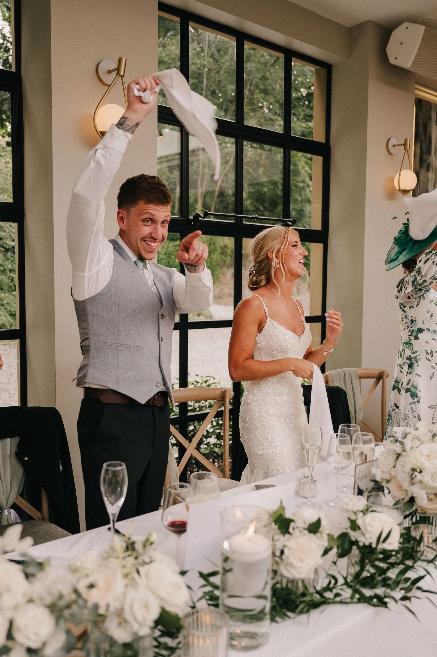Joyful couple celebrates at wedding reception with elegant white floral table decor and candlelight in sunlit venue.