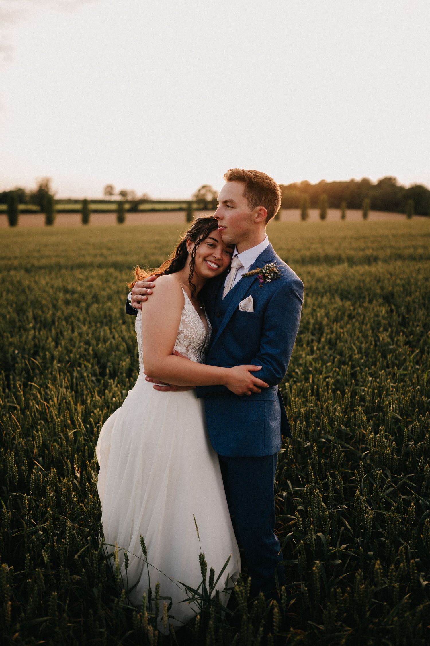 A romantic wedding couple embraces in a golden field at sunset wearing a white dress and navy suit.