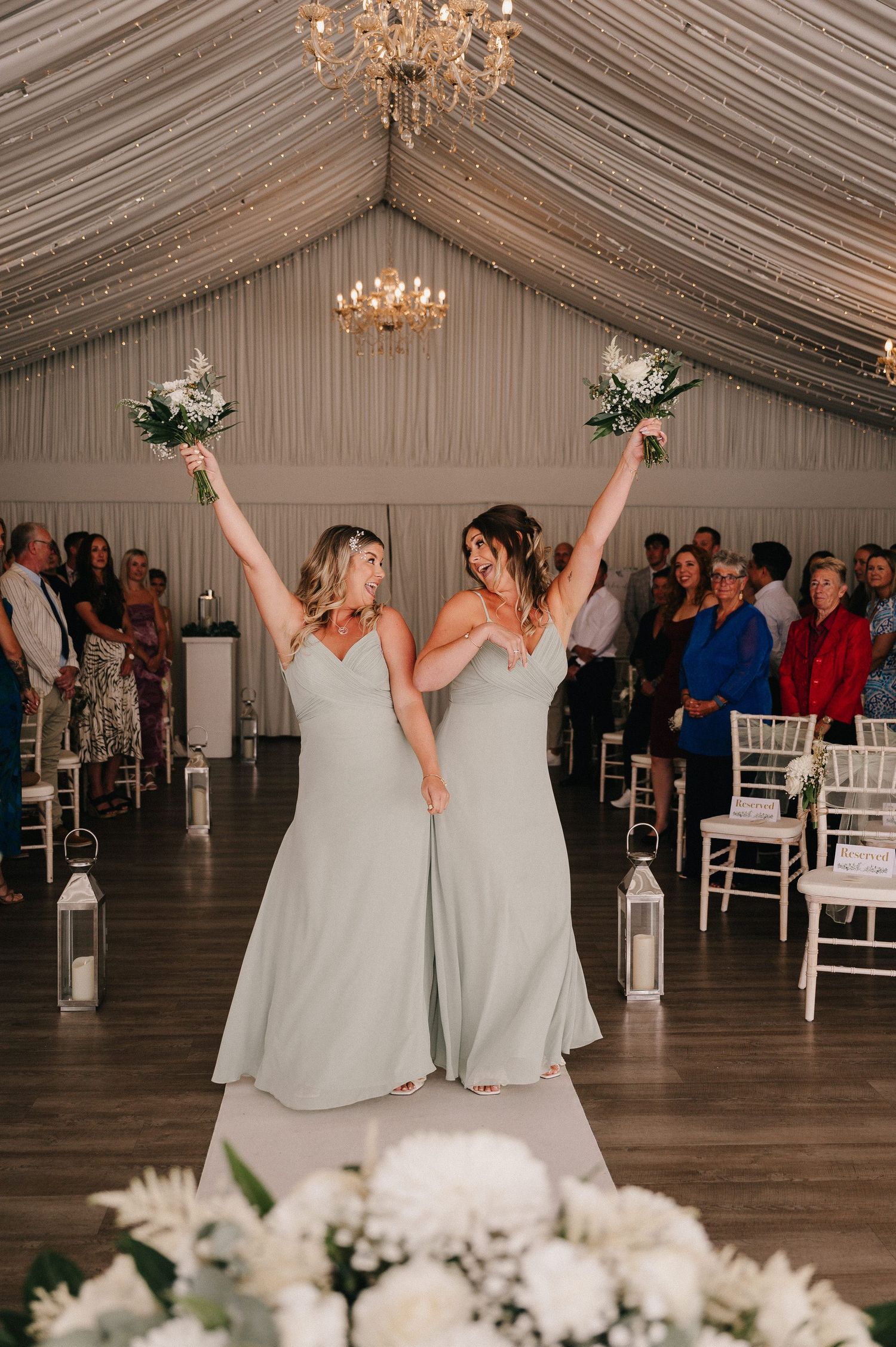 Two people in white dresses dancing and celebrating together in an elegant tent reception venue with chandeliers.