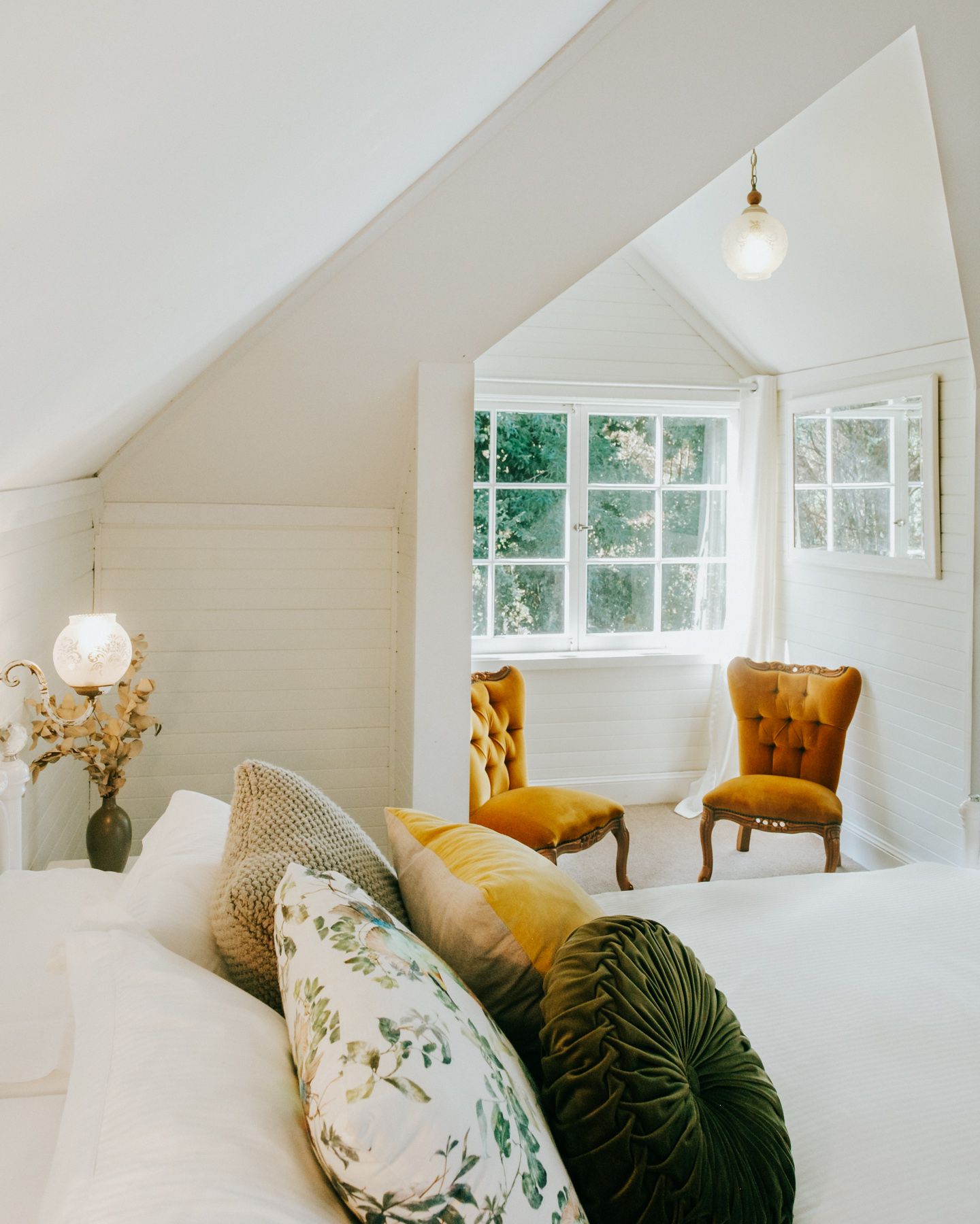 Bright white attic bedroom with vaulted ceiling features yellow velvet chairs and floral accent pillows by windows.