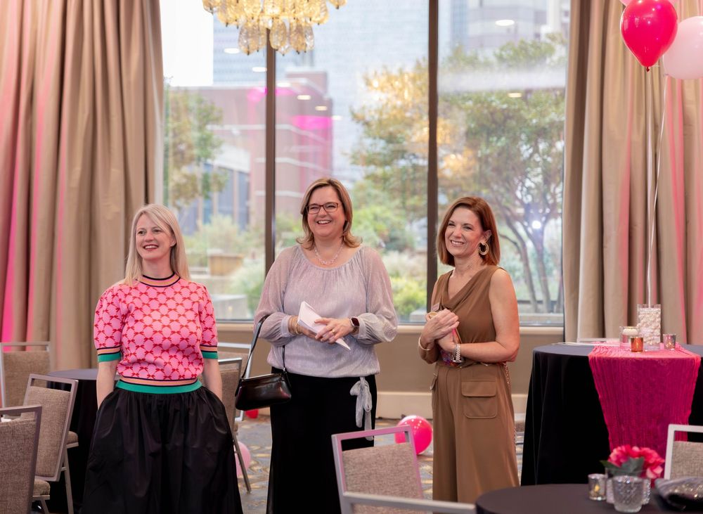 Three women stand together at an elegant event space with a chandelier and pink decor.