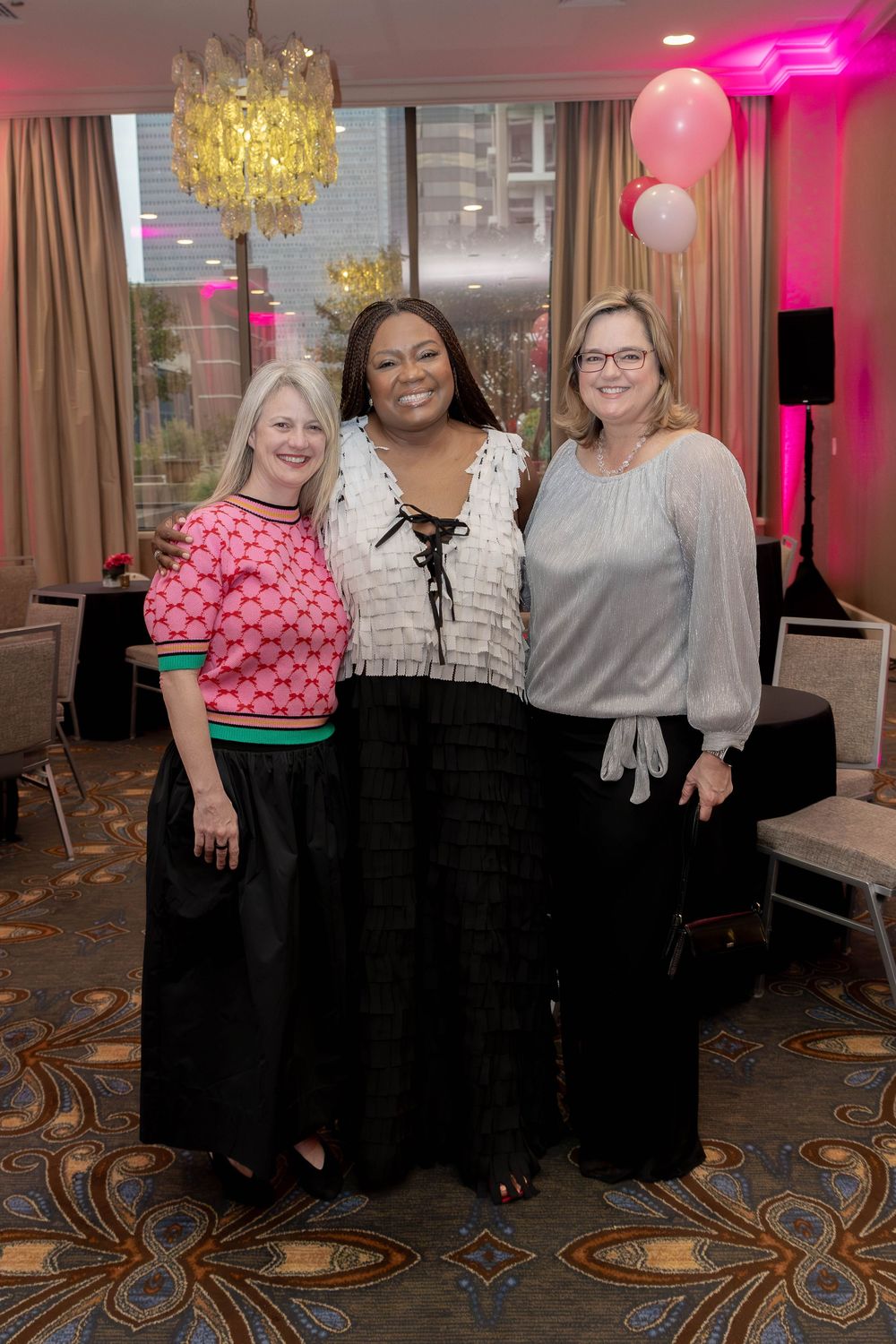 Three women pose together under a golden chandelier at an elegant event with pink lighting.