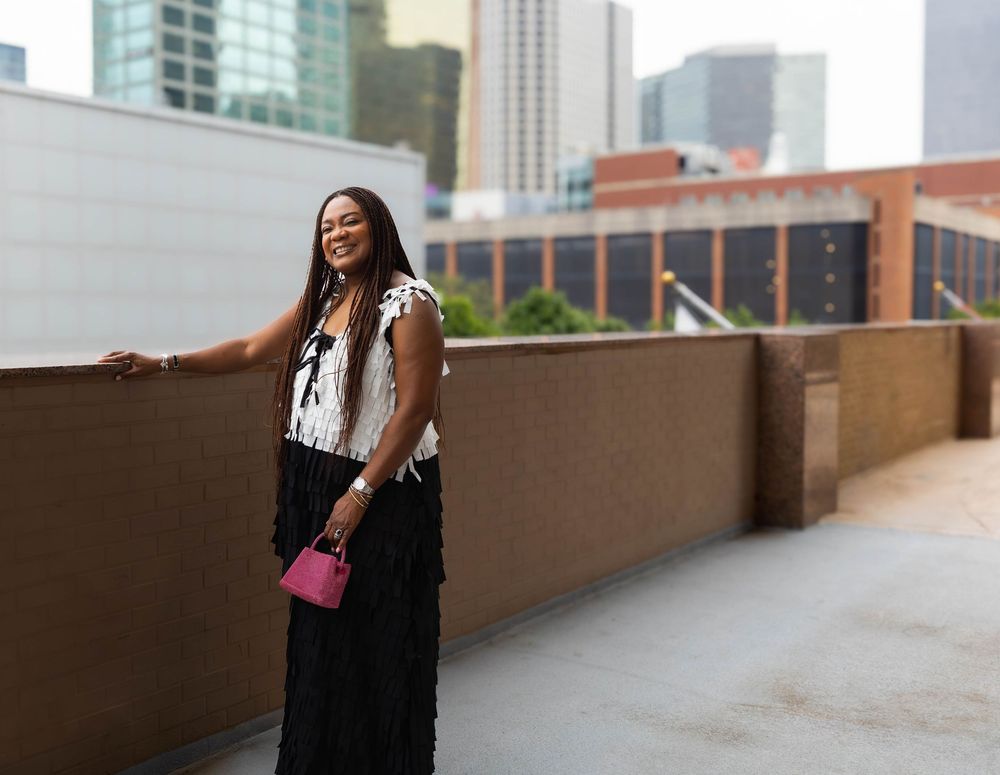 Woman in black and white outfit poses on urban rooftop terrace with city buildings in background.