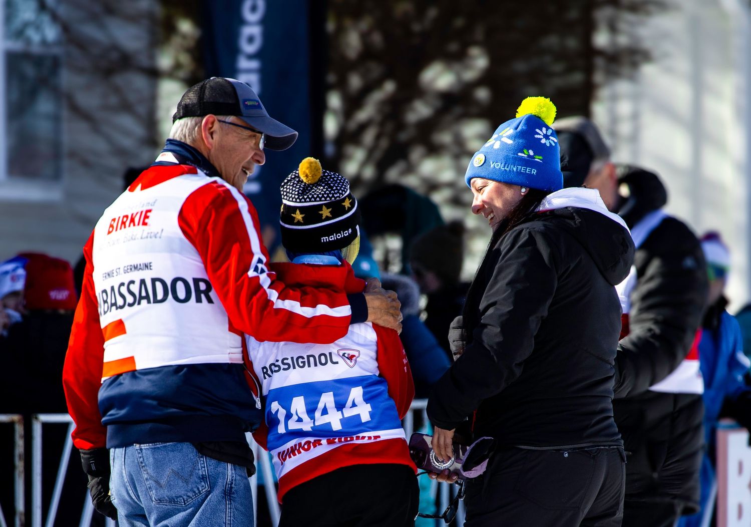 A group of skiers gather at a snowy ski resort with some wearing red and blue jackets while others watch nearby.