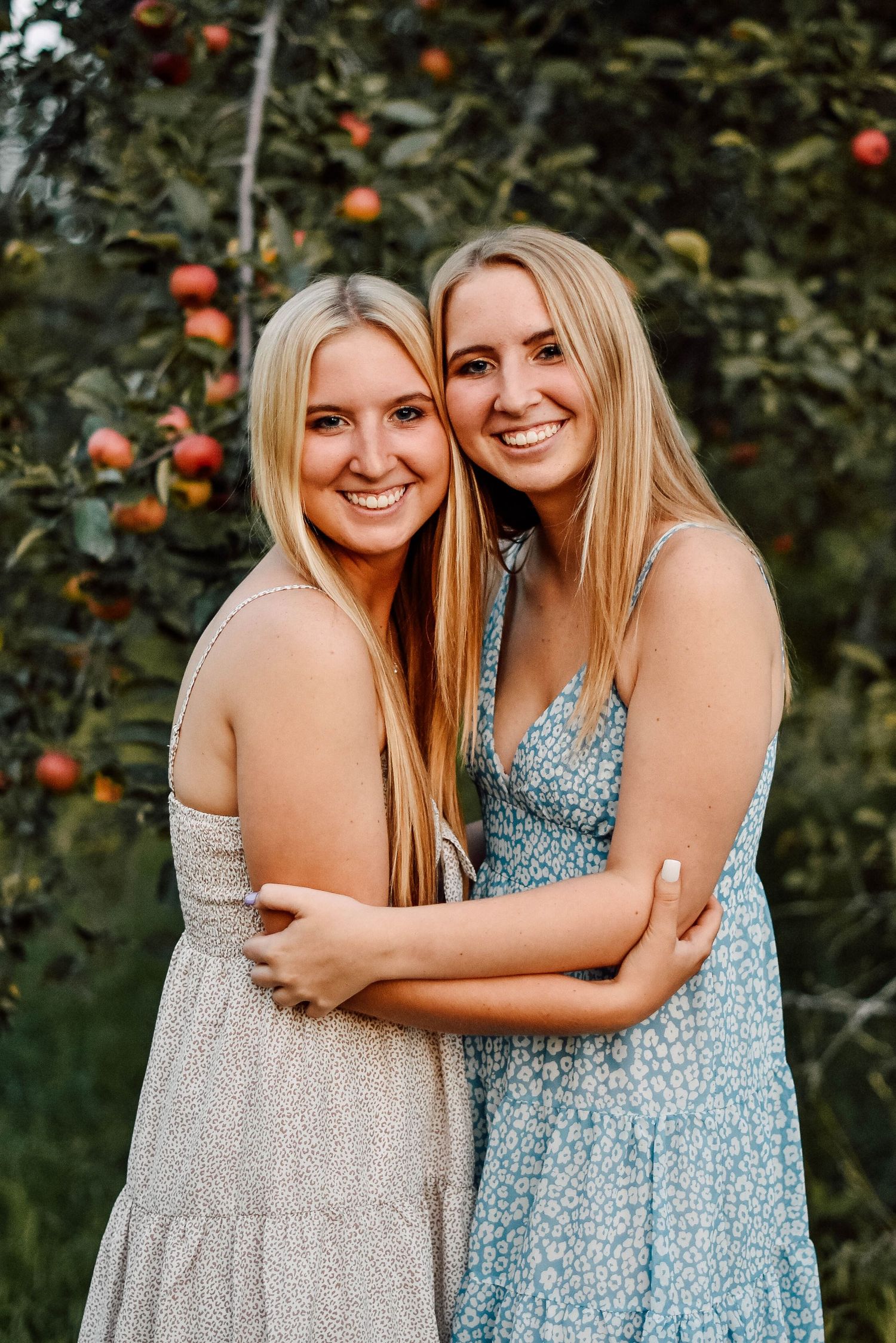 Friends in summer dresses embrace in front of orange tree.